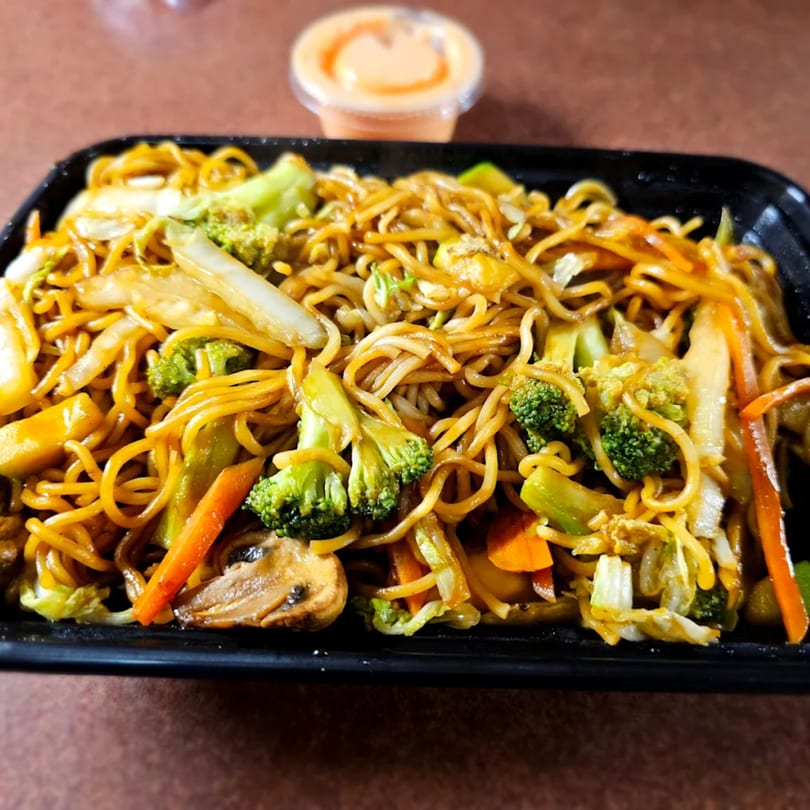 Close-up of lo mein noodles with broccoli, carrots, cabbage, and mushrooms in a black container.