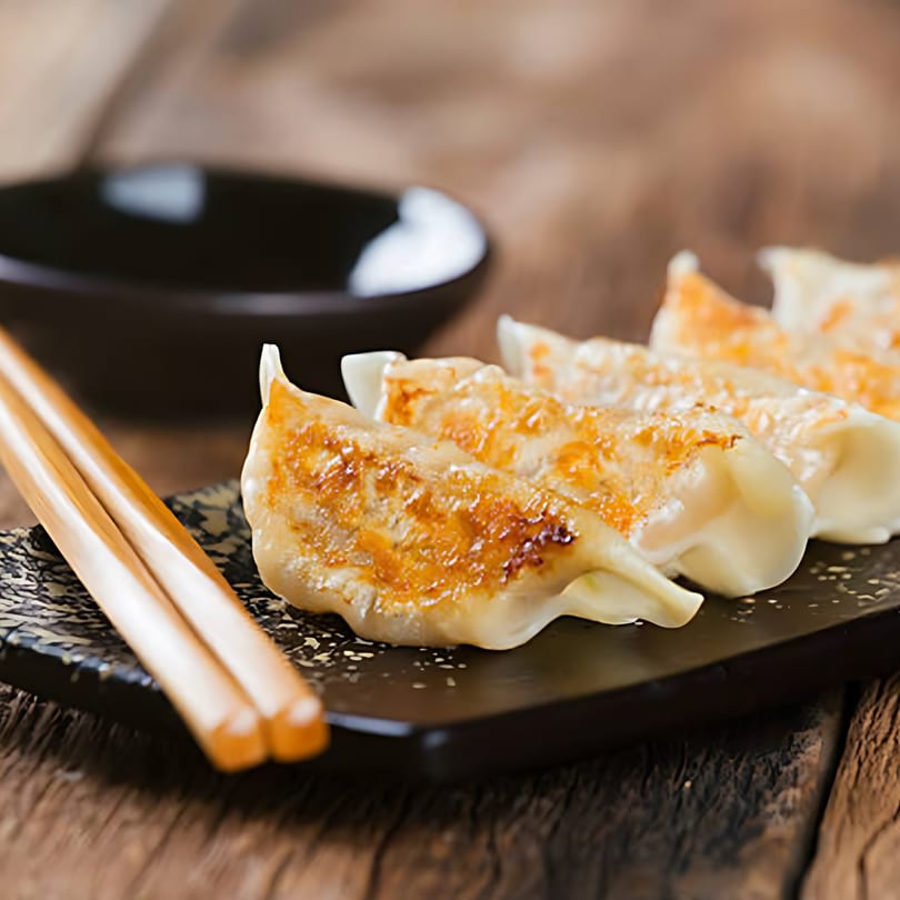 Golden-brown pan-fried gyoza dumplings on a black plate with chopsticks and a dipping bowl.