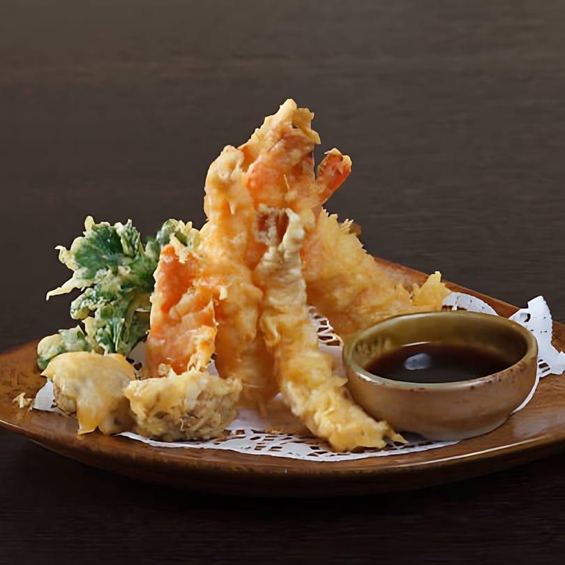 Crispy shrimp and vegetable tempura with a side of dipping sauce on a wooden plate.