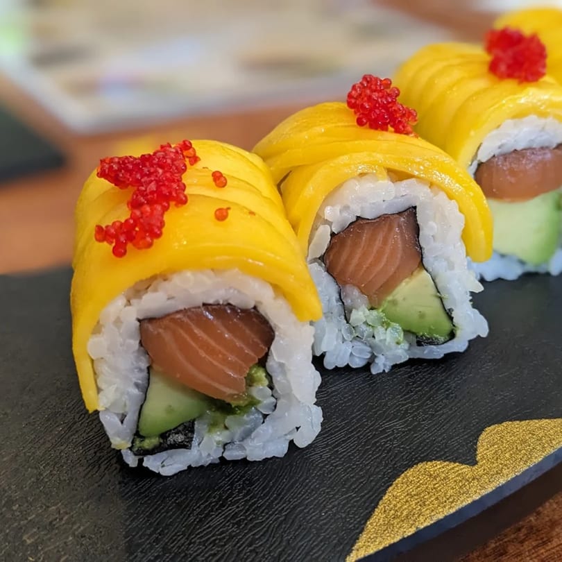 Three mango sushi rolls with salmon, avocado, and red roe on a black plate.